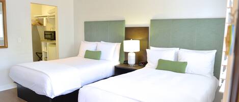 A hotel room with two beds, each with white bedding and green pillows, a bedside table with a lamp, and a small TV.
