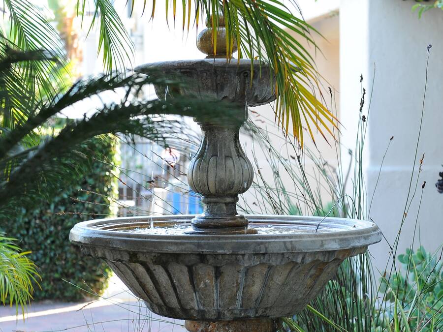 A stone fountain with a tiered design, surrounded by lush greenery and a palm tree.
