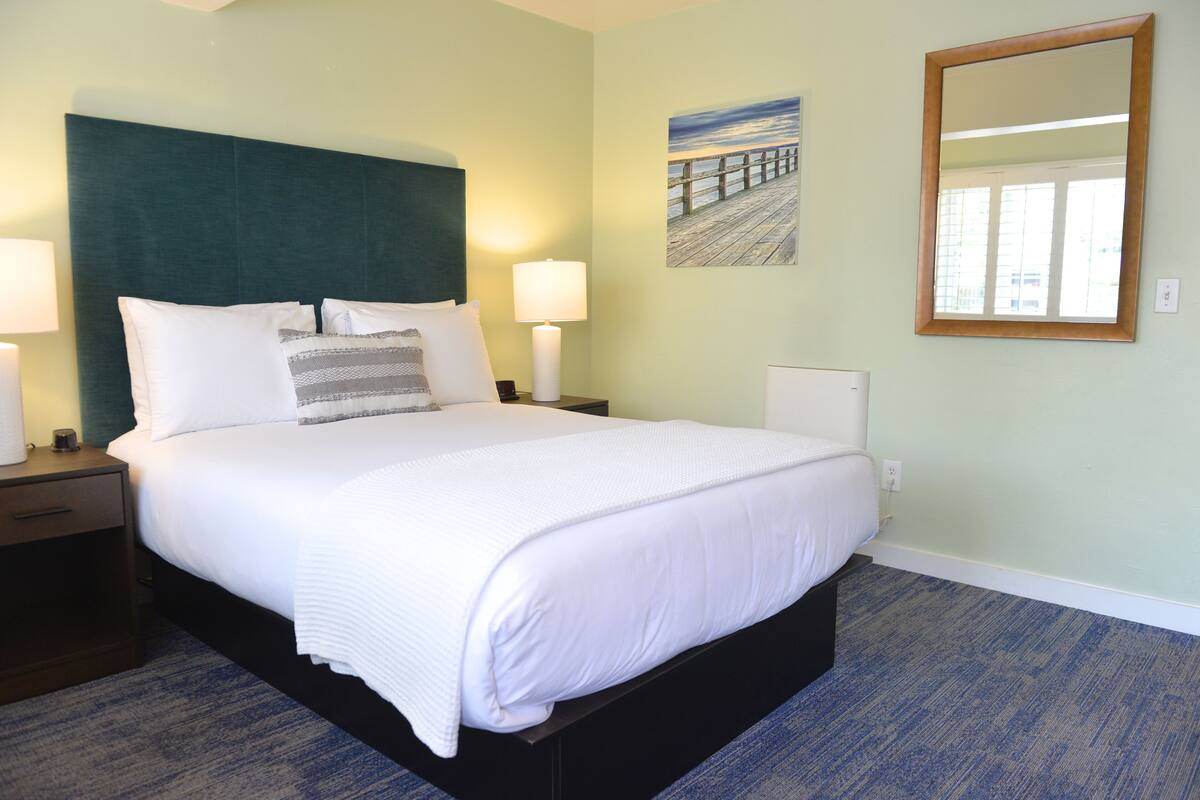 A neatly made bed with white linens and a dark headboard, a bedside lamp, and a painting of a pier on the wall.