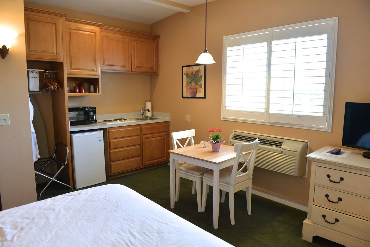 A compact hotel room with a bed, a small kitchenette, a dining area with a table and chairs, a TV, and a window with blinds.