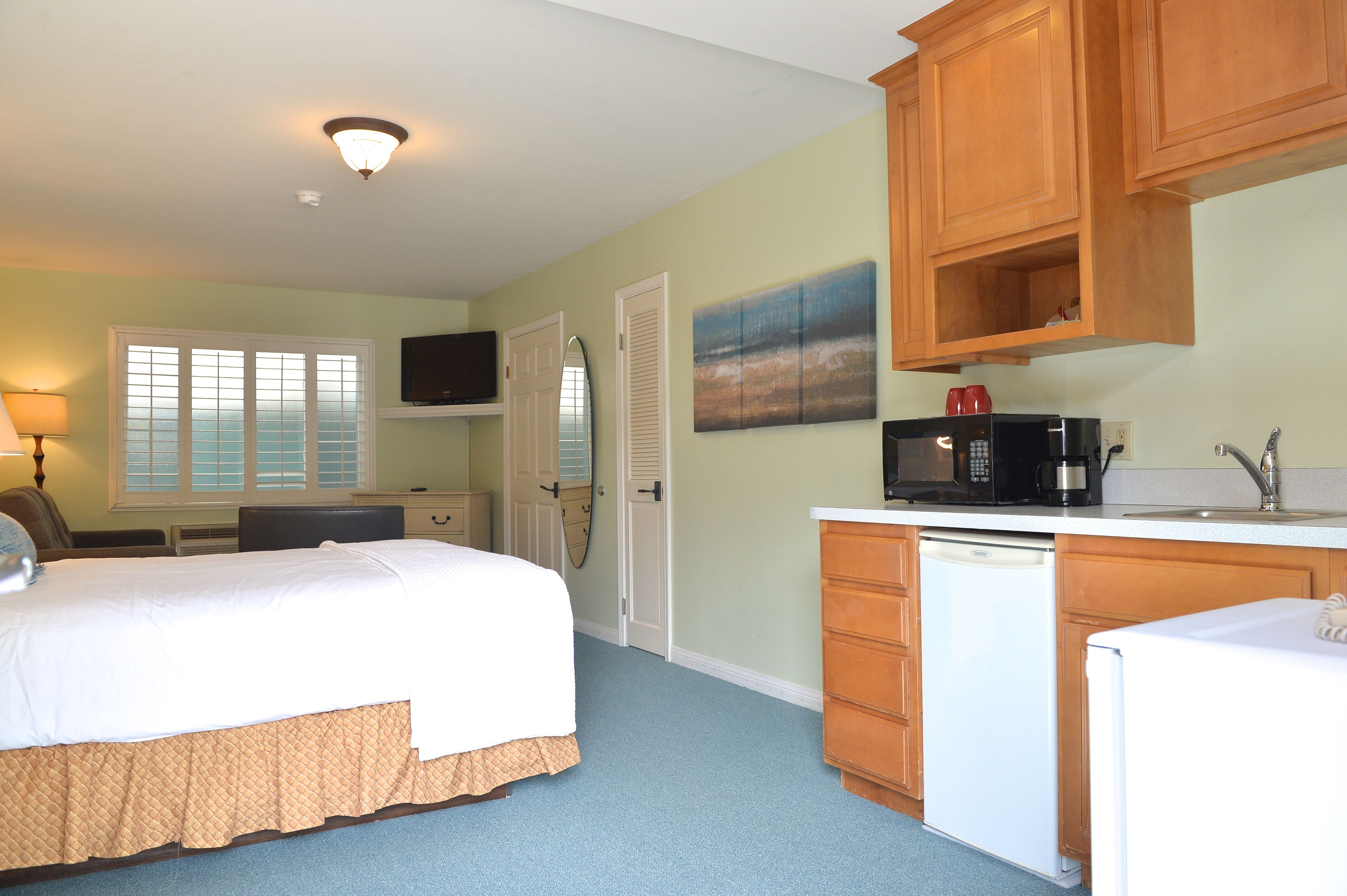 A bedroom with a bed, a desk, a chair, a television, and a kitchen area with a microwave, sink, and cabinets.