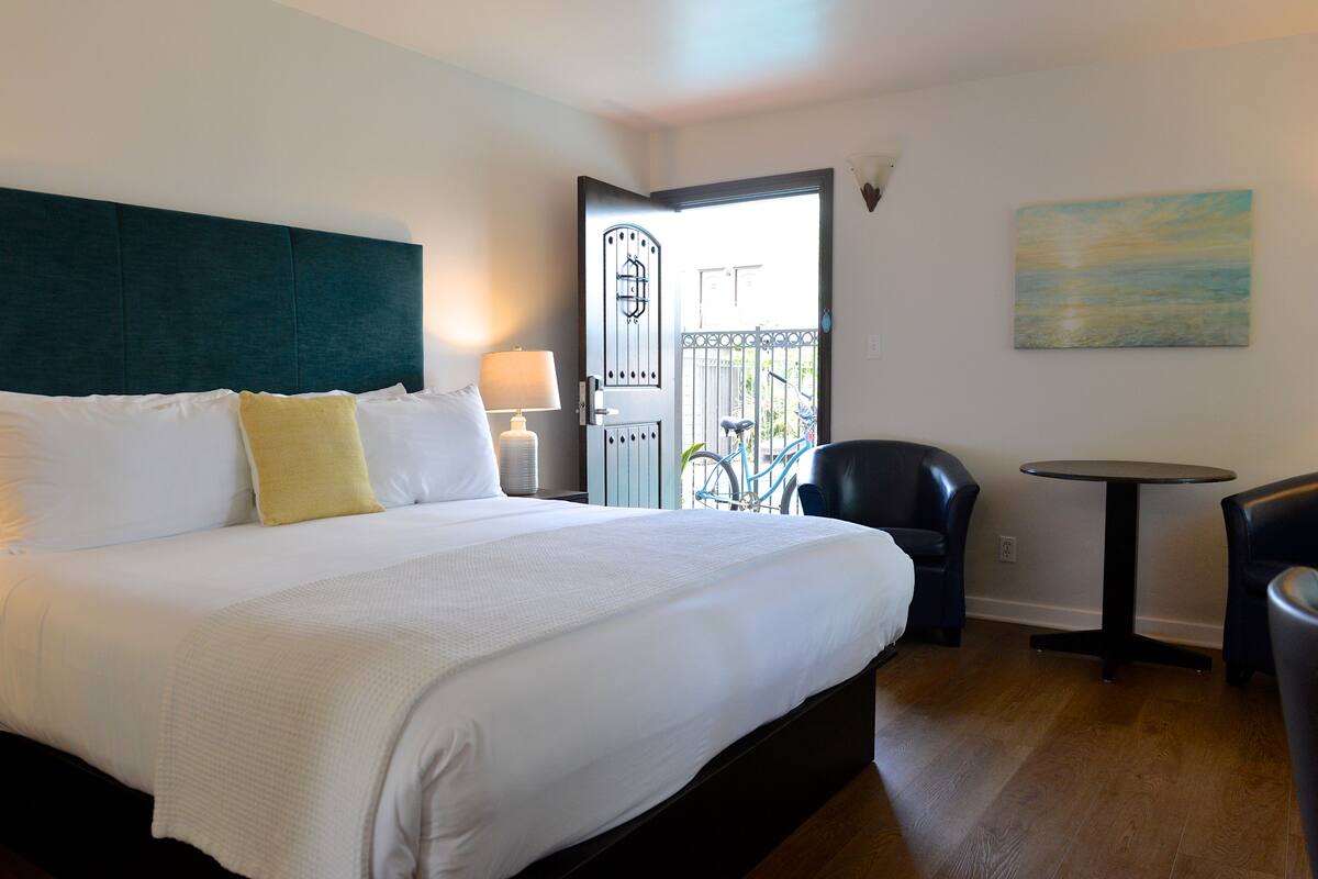 A hotel room with a large bed, a desk, a chair, a balcony, and a painting on the wall.