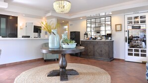 Een receptiezone met een ronde tafel, vazen en een kroonluchter. Achterin staat een balie met een computer en een koelkast.