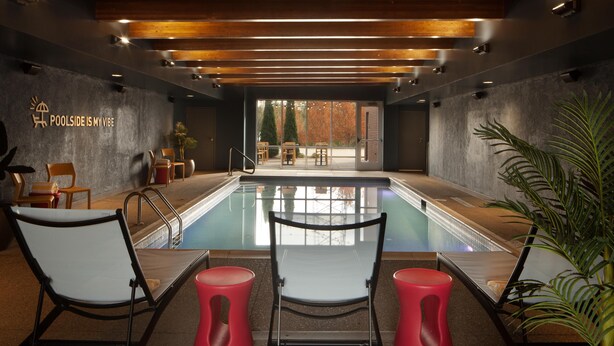 Un espace piscine intérieur moderne avec vue sur l’extérieur, des coins salon et un panneau indiquant : « J’ADORE L’AMBIANCE DE LA PISCINE ».