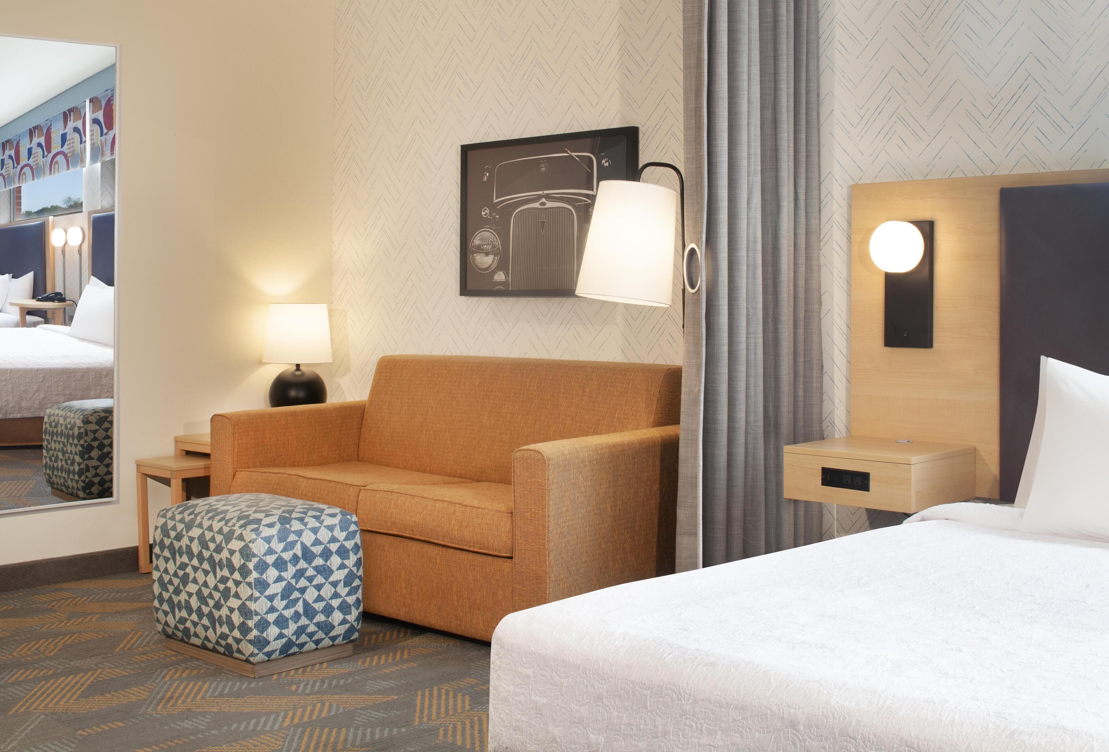 A hotel room with a bed, a sofa, a small ottoman, a nightstand, a lamp, a wall-mounted light, and a framed picture.