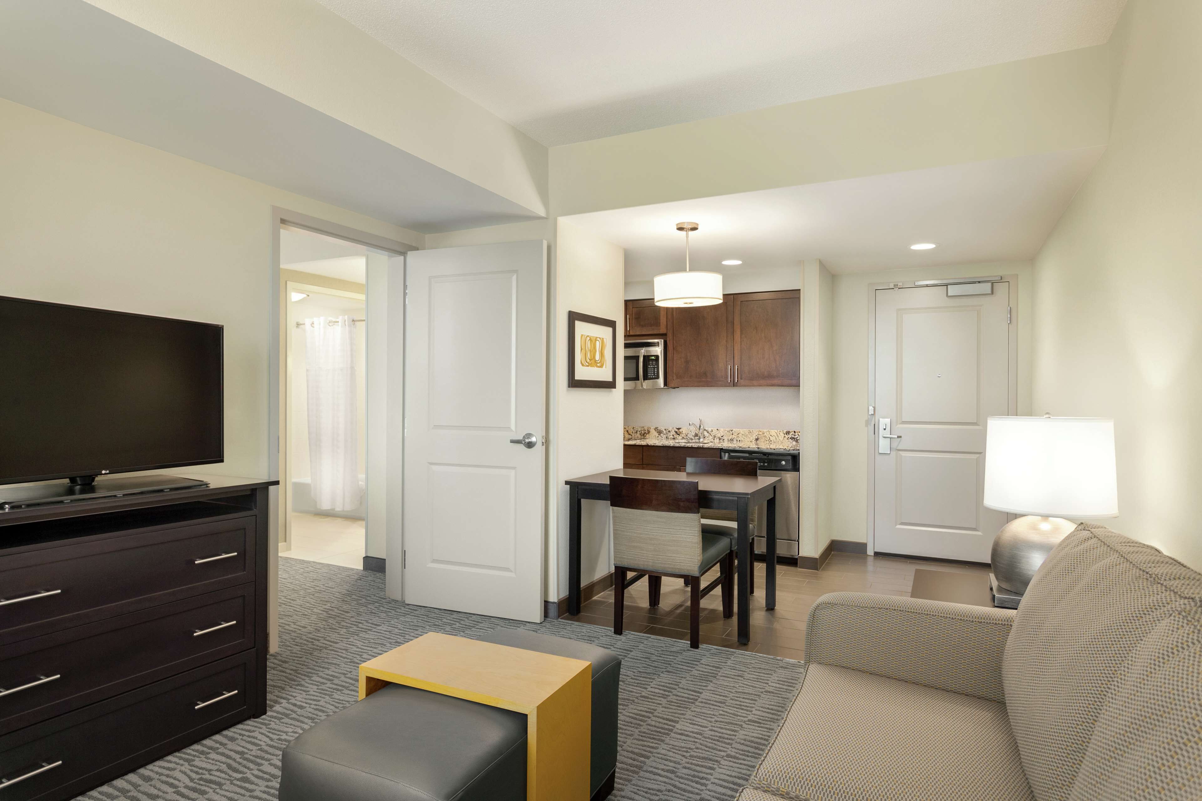 A hotel room with a flat-screen TV, a dark wooden dresser, a beige sofa, a small wooden coffee table, and a dining area with a kitchen in the background.