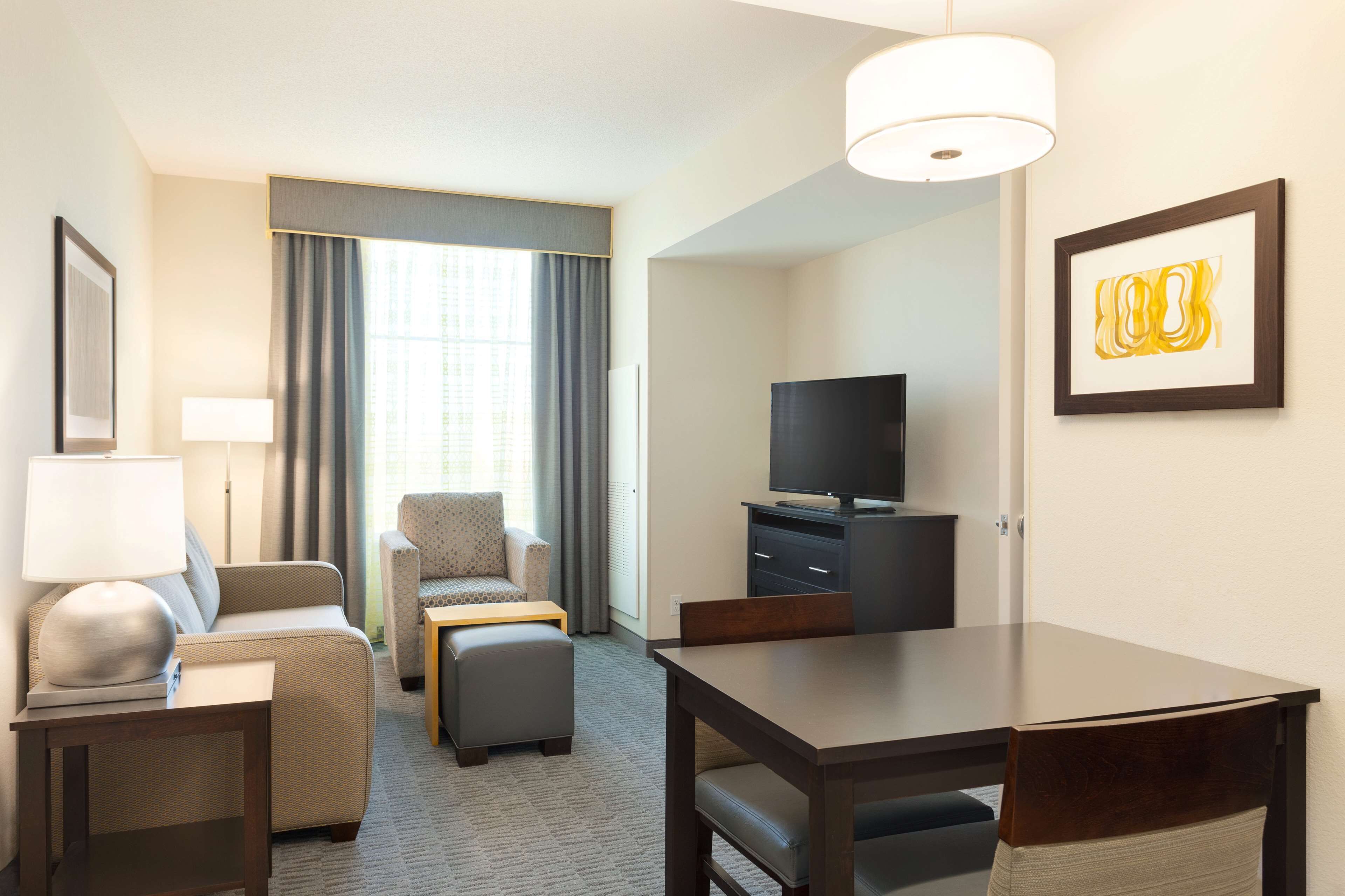 Ein Hotelzimmer mit Sofa, Sessel, Hocker, Esstisch, Fernseher und Kunstwerk an der Wand.