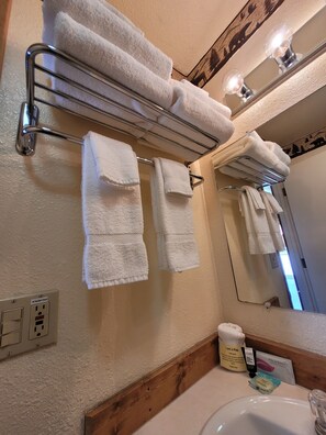 A bathroom with a towel rack holding several folded towels, a mirror, a sink, and toiletries on the counter.