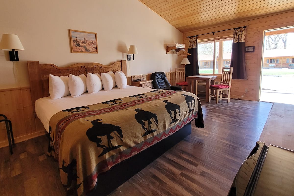 A bedroom with a large bed, wooden headboard, and a blanket with horse silhouettes. There are two bedside tables with lamps, a chair, and a desk with a chair.