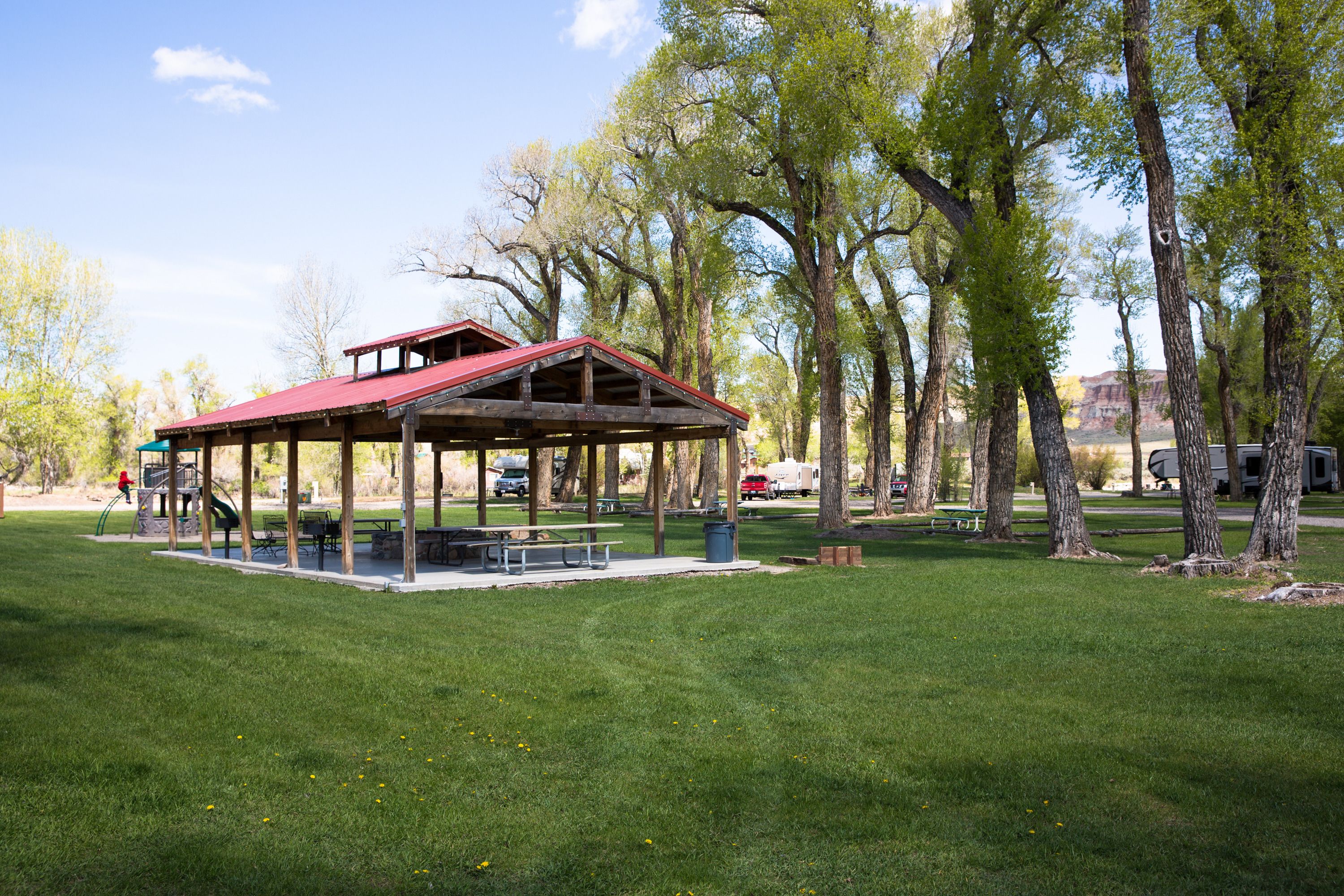 A covered picnic area with benches and tables, surrounded by trees and open grassy space.