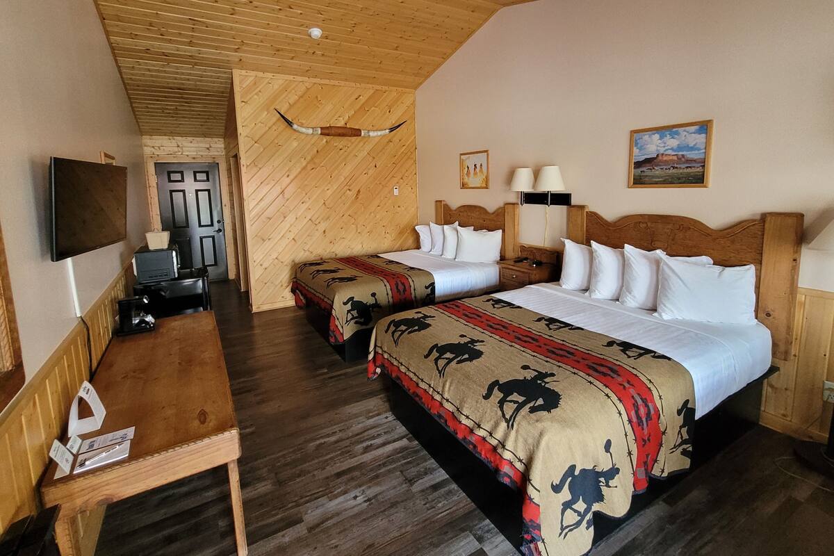 A hotel room with two beds, wooden floors, a wooden ceiling, a mounted TV, a desk, and a door.