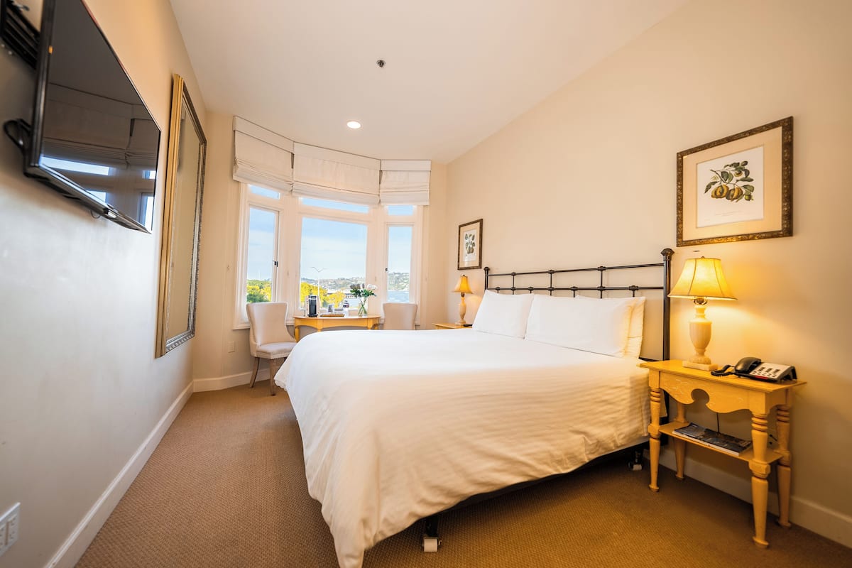 A hotel room with a large bed, a bedside table with a lamp, a chair, a TV mounted on the wall, and a window with a view.