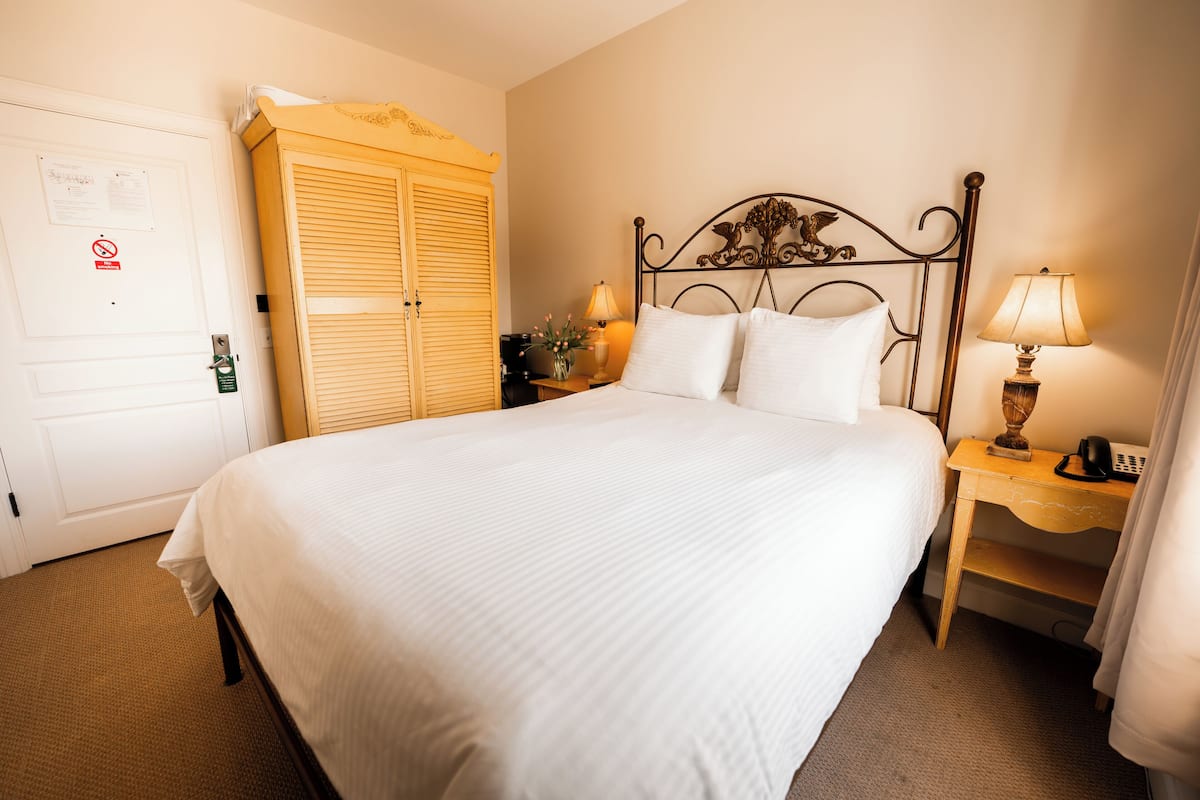 A neatly made bed with white linens, a wooden wardrobe, a bedside table with a lamp, and a nightstand with a phone.