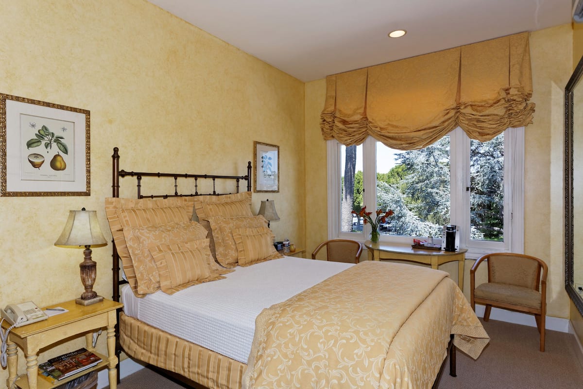 A bedroom with a large bed, a bedside table with a lamp, a chair, and a window with curtains.