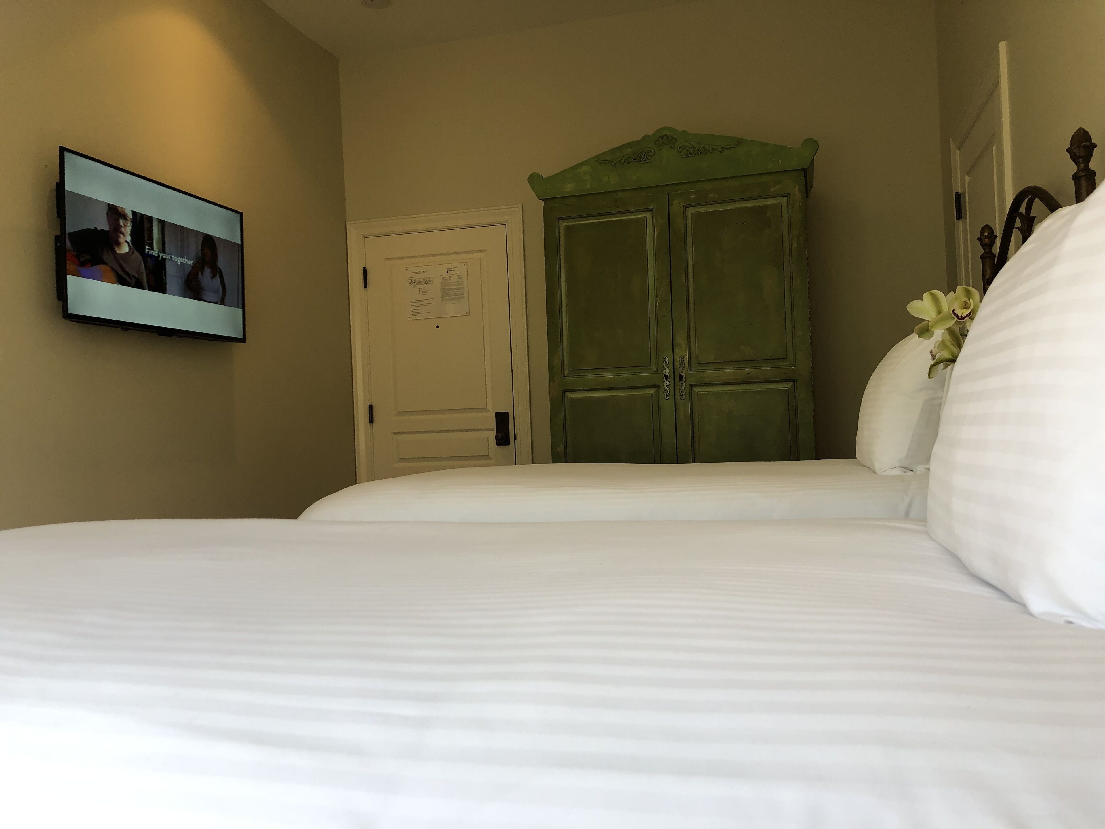 Una habitación de hotel con una cama, un armario verde, una puerta con un aviso y un televisor mostrando una escena.