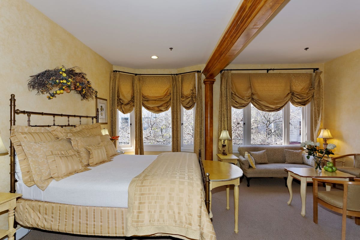 A bedroom with a large bed, a sitting area with a sofa and a table, and a view of trees through the windows.