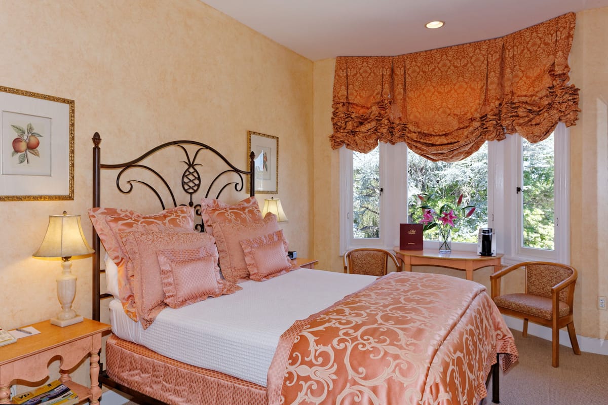 A bedroom with a large bed, a nightstand with a lamp, a chair, and a window with curtains.