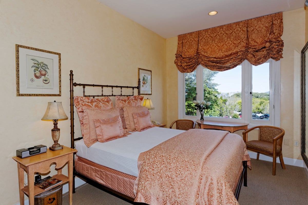 A bedroom with a bed, bedside table, lamp, chair, and a window with a view of greenery.