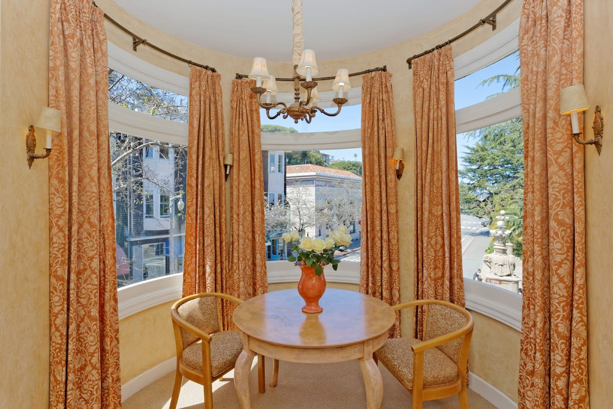 A corner room with a round table, two chairs, a vase with flowers, a chandelier, and curtains.