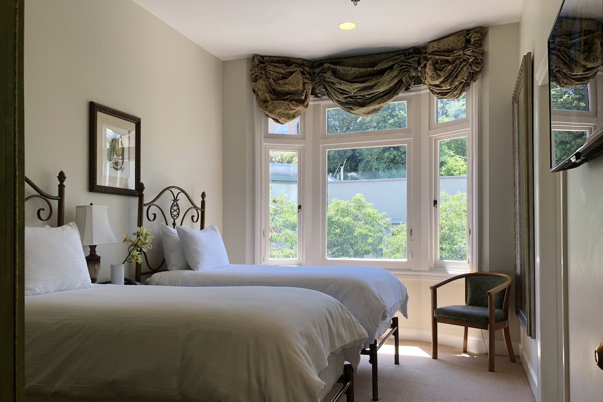 A bedroom with two beds, a chair, a window with curtains, a framed picture, and a lamp.