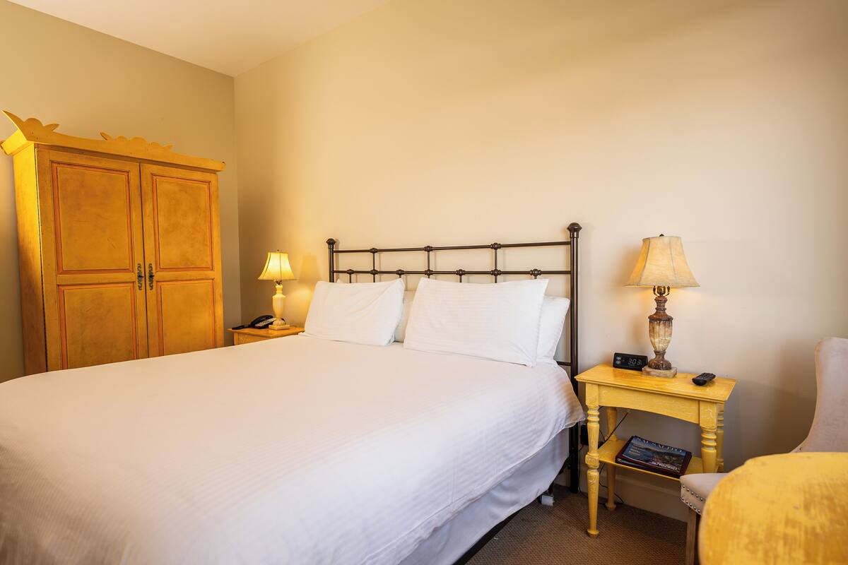 A neatly made bed with white linens, a wooden wardrobe, and two bedside tables with lamps and a book.