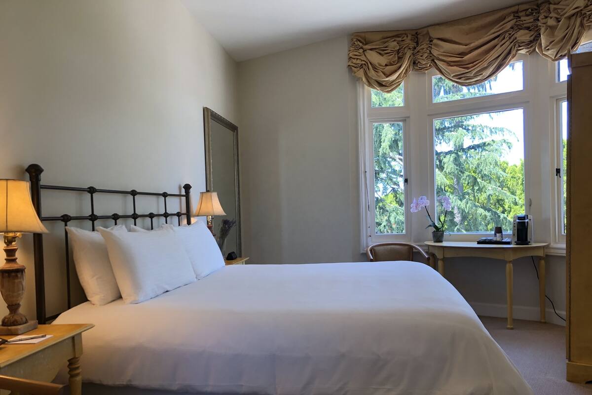 A neatly made bed with white linens, a bedside lamp, a small table with a coffee maker, and a chair.