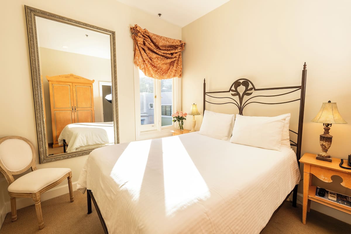A neatly made bed with white linens, a large mirror, a wooden chair, and a bedside table with a lamp.