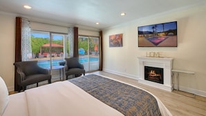 A hotel room with a large bed, two armchairs, a fireplace, and a view of a pool and outdoor area. - Little Paradise Hotel (Palm Springs)