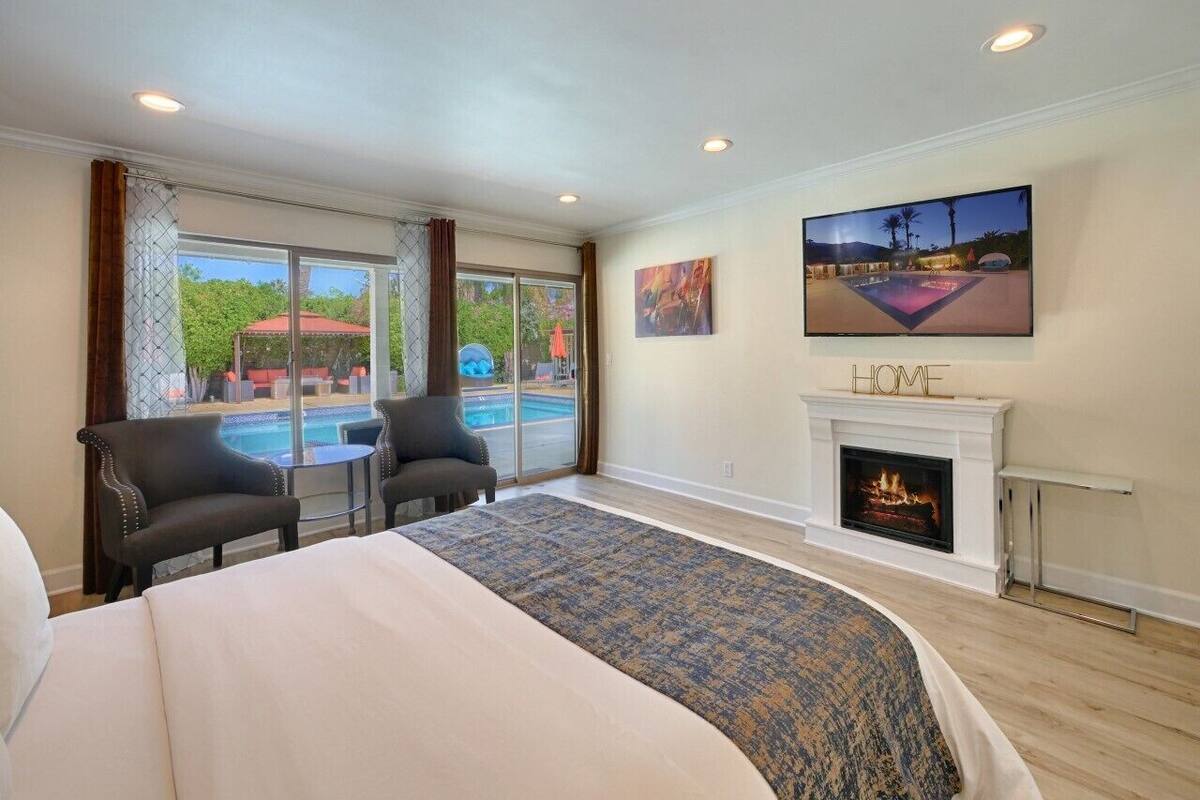 A hotel room with a large bed, two armchairs, a fireplace, and a view of a pool and outdoor area.