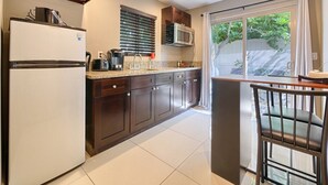A modern kitchen with dark wood cabinets, a white refrigerator, a microwave, and a sink. There is a window with blinds and a view of greenery outside. - Little Paradise Hotel (Palm Springs)