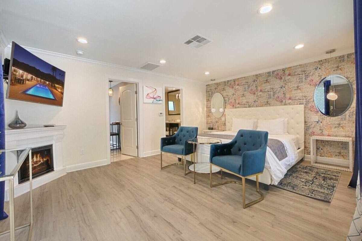 A modern bedroom with a fireplace, a bed, two blue armchairs, a round mirror, and a television displaying a pool scene.