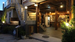 Un bâtiment à plusieurs niveaux, avec un escalier menant à un étage supérieur, un espace bar doté d’un comptoir et d’étagères, et un petit espace extérieur aménagé avec des plantes.