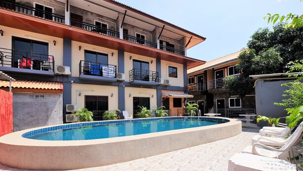 Una piscina con sillas de descanso, un edificio de varios pisos con balcones y una zona de patio con baldosas.