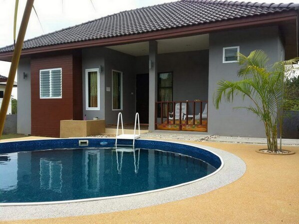 A residential swimming pool with a tiled roof, a covered patio, and a small garden area with palm trees.
