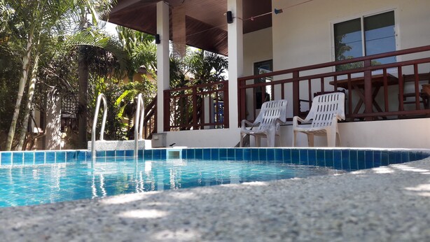 A swimming pool with a tiled edge, surrounded by white plastic chairs, and a covered area with a wooden roof. - Lanta Dream Garden Pool Villa (Ko Lanta)