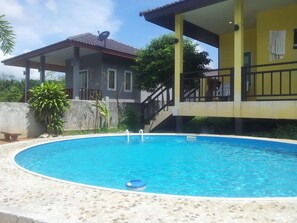 Una piscina con borde de piedras, una zona techada y una casa con antena parabólica.
