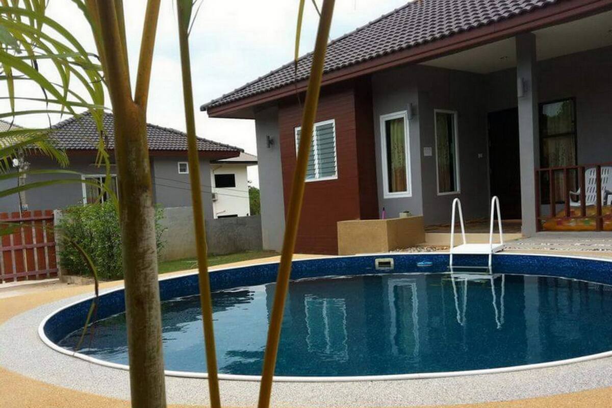 A swimming pool with a tiled deck, surrounded by a house with a tiled roof and a fenced yard.