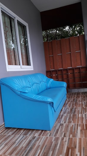 Un sofá azul en una habitación con piso estampado, una ventana con cortinas florales y una valla de madera en el exterior.