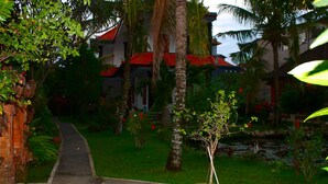 A pathway leading to a building with a red roof, surrounded by lush greenery and palm trees. - Swan Inn (Ubud)