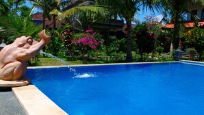 A swimming pool with a frog statue spouting water, surrounded by lush greenery and a building with a red-tiled roof. - Swan Inn (Ubud)