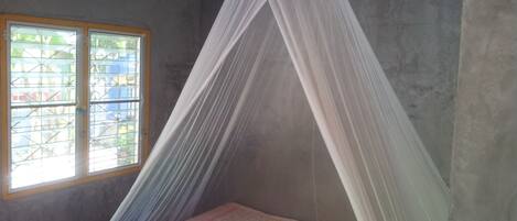 A room with a bed under a white mosquito net, a window with bars, and a concrete wall.