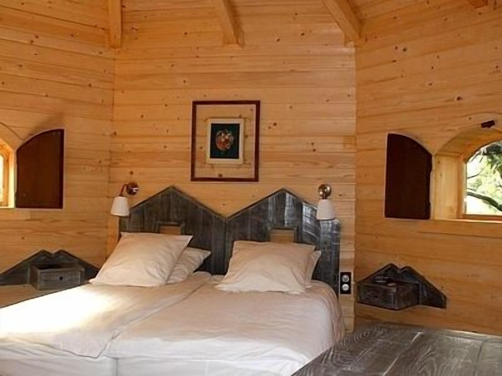 A wooden cabin bedroom with a bed, two bedside lamps, a framed picture on the wall, and a window with a view of greenery.