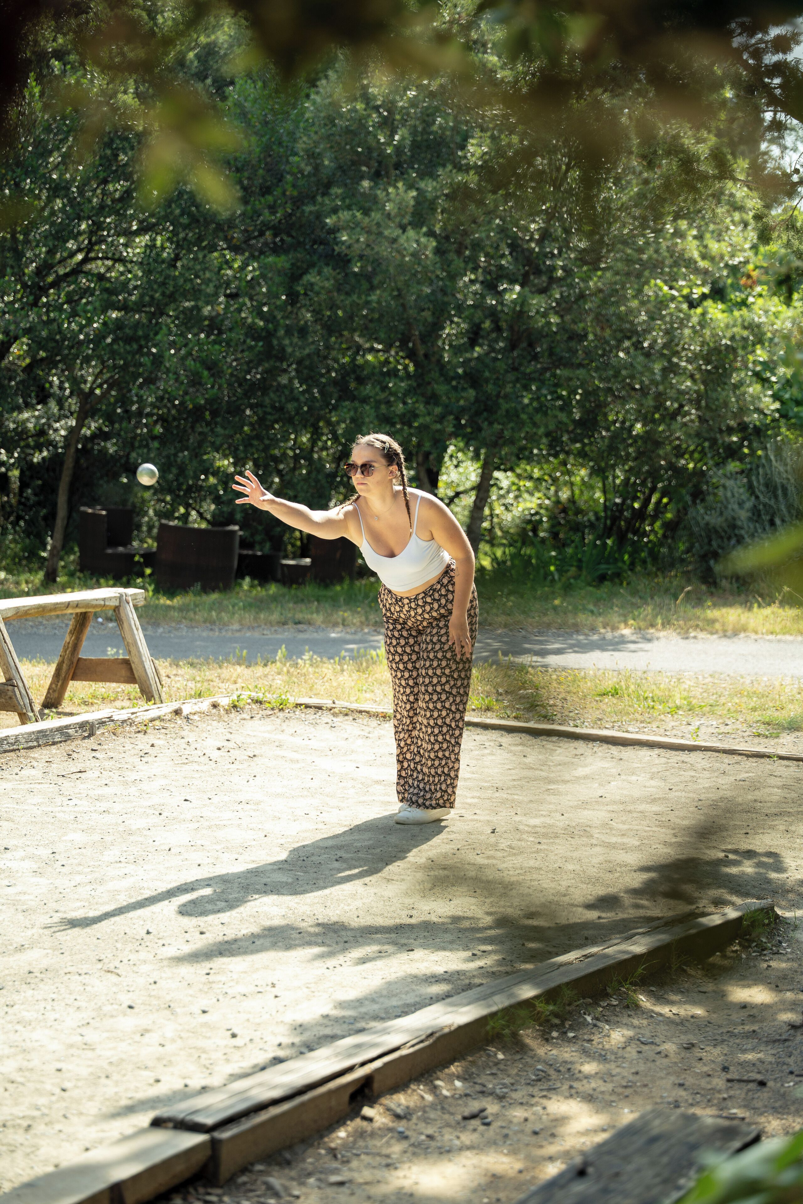A person in a white tank top and patterned pants standing on a concrete path with a wooden structure in the background.