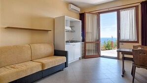 A room with a sofa, a small kitchenette, a dining table, and a view of the sea through a glass door.