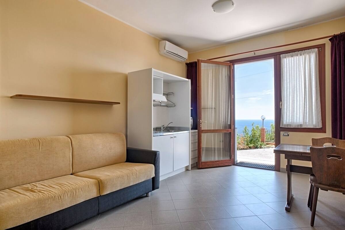 Une pièce comprenant un canapé, un petit coin cuisine, une table à manger et donnant sur la mer grâce à une porte vitrée.