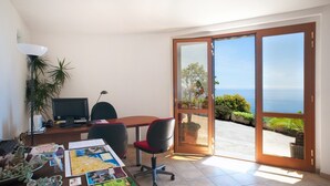 A room with a desk, computer, and office chair, a potted plant, and a view of the ocean through a sliding glass door.