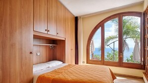 A bedroom with a bed, wooden wardrobe, and a large window with a view of palm trees and the sea.
