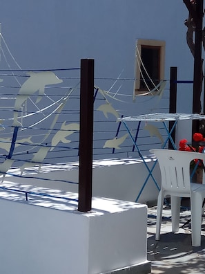 A white outdoor seating area with a blue and white striped fence, a small window, and a white plastic chair. - Villa Diktynna (Hersonissos)