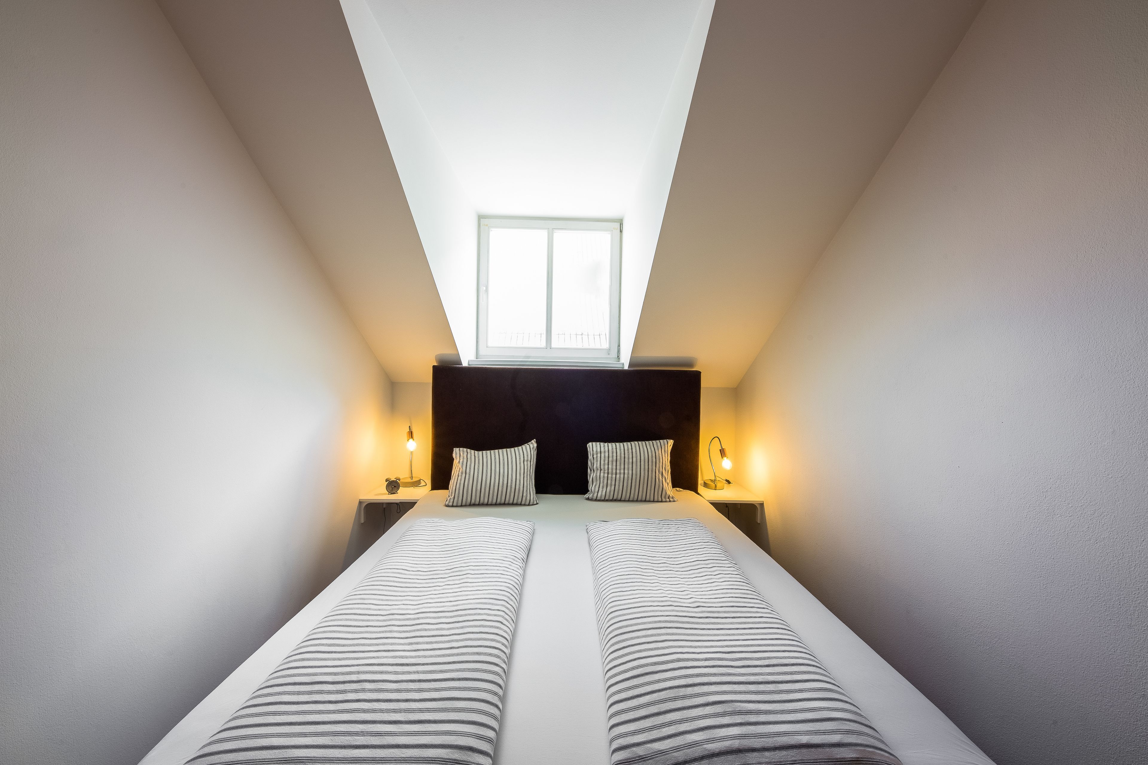 Une chambre avec un plafond en pente, un puits de lumière, un lit avec une literie à rayures, deux tables de chevet avec des lampes et une tête de lit sombre.