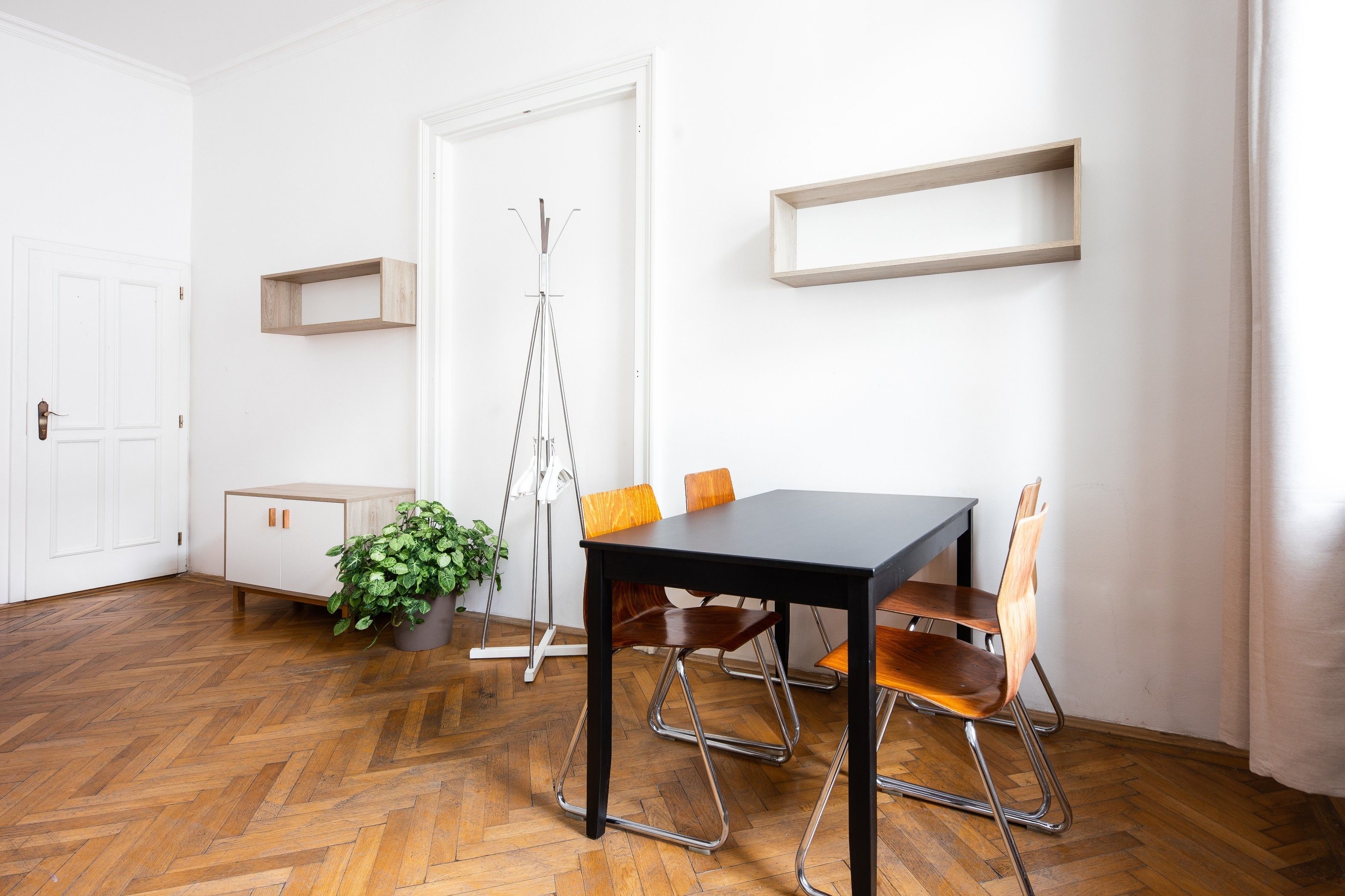 Ein Zimmer mit Holzboden im Fischgrätmuster, einem schwarzen Tisch mit vier Stühlen, einem Blumentopf, einer Stativlampe und einer weißen Tür mit kleinem Schrank.
